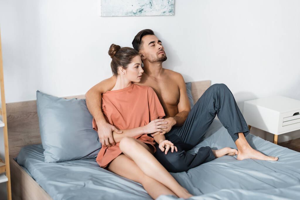 muskularny mężczyzna w piżamie spodnie siedzi na łóżku z zamkniętymi oczami i trzyma się za ręce z dziewczyną w t-shircie - Zdjęcie, obraz