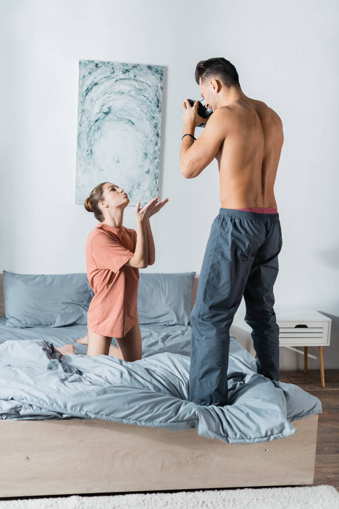 sexy vrouw in t-shirt knielen op bed en blazen lucht kus in de buurt shirtless vriendje met vintage camera - Foto, afbeelding