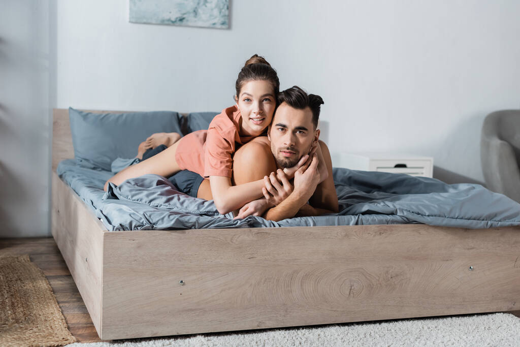 junges und sexy Paar lächelt in die Kamera und umarmt sich auf grauer Bettwäsche - Foto, Bild