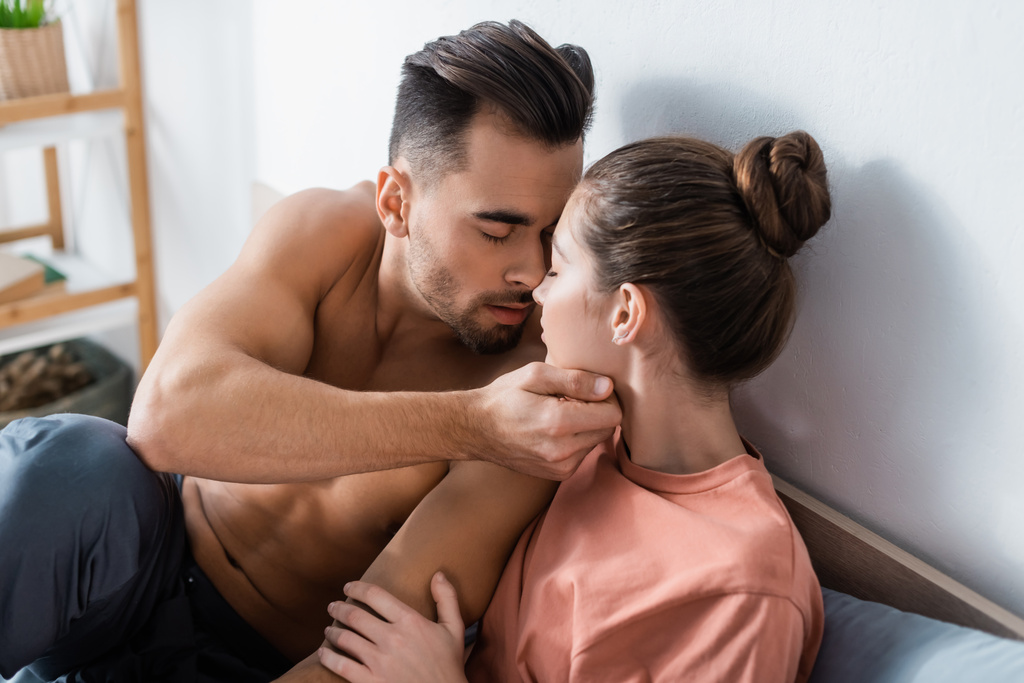 sexy muscular man with closed eyes touching face of girlfriend in bedroom - Photo, Image