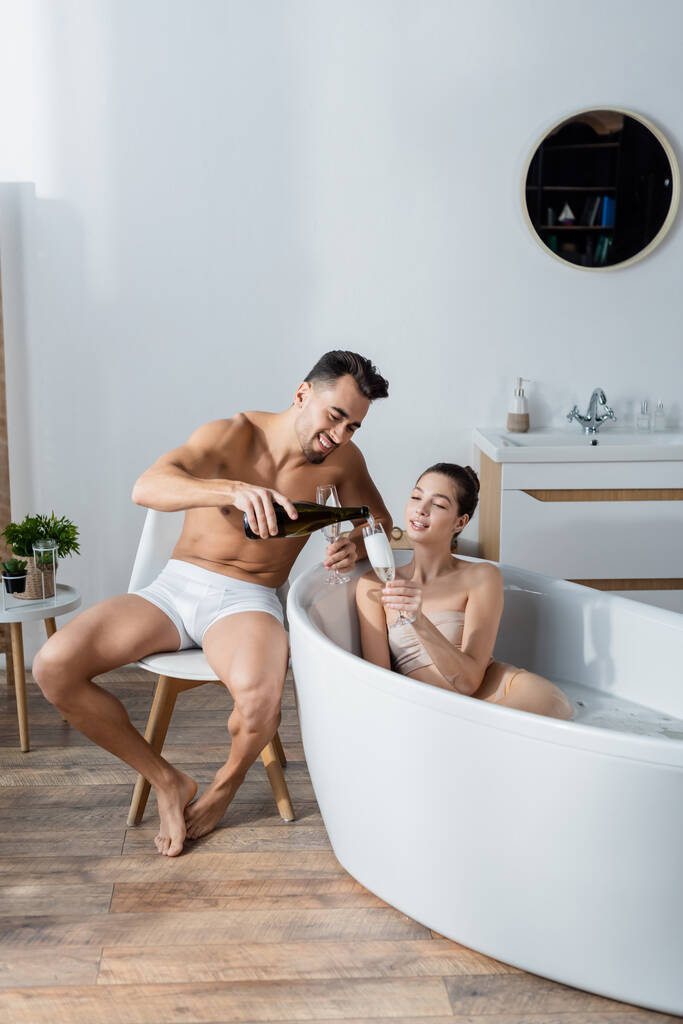 glücklicher Mann in Unterhosen, der Champagner ins Glas gießt, Frau entspannt sich in Badewanne - Foto, Bild