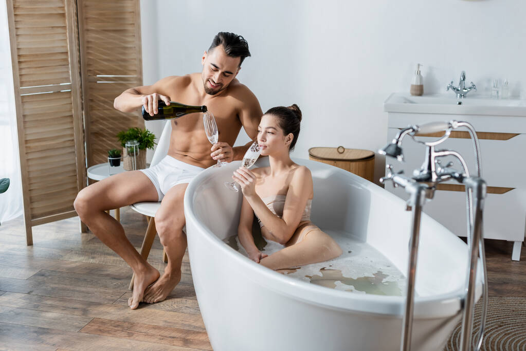 hymyilevä mies alushousut kaatamalla samppanjaa lähellä tyttöystävä kylvyssä tilava kylpyhuone - Valokuva, kuva