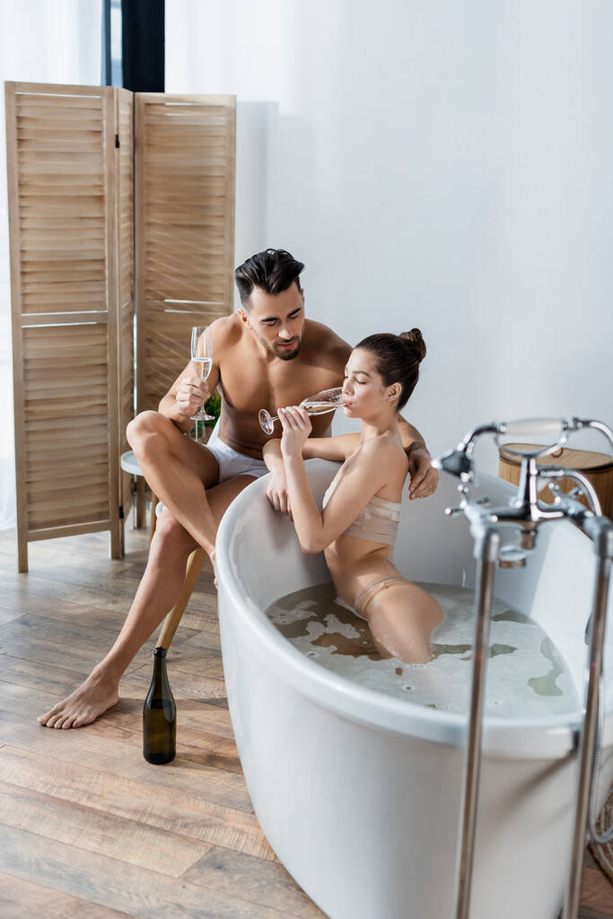 seductora mujer bebiendo champán en bañera cerca de novio en calzoncillos - Foto, Imagen