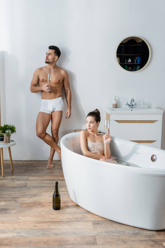 homme sexy en caleçon debout avec verre de champagne près de petite amie se détendre dans la baignoire - Photo, image