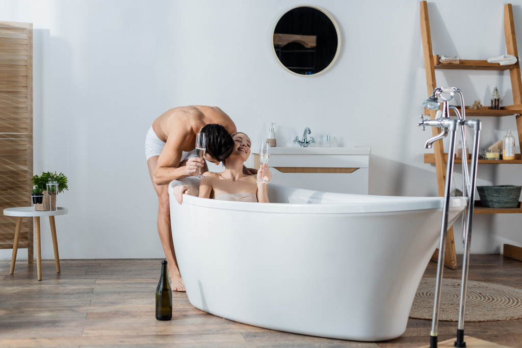 молодий чоловік тримає келих шампанського і цілує усміхнену дівчину, що розслабляється у ванній
 - Фото, зображення
