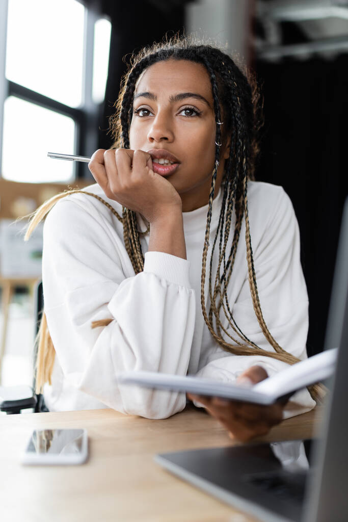 Femme d'affaires afro-américaine pensive tenant un stylo et un carnet près de gadgets flous  - Photo, image