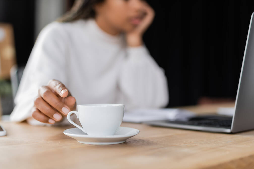 Ausgeschnittene Ansicht einer afrikanisch-amerikanischen Geschäftsfrau, die eine Tasse Kaffee in der Nähe von Laptop auf dem Tisch nimmt  - Foto, Bild