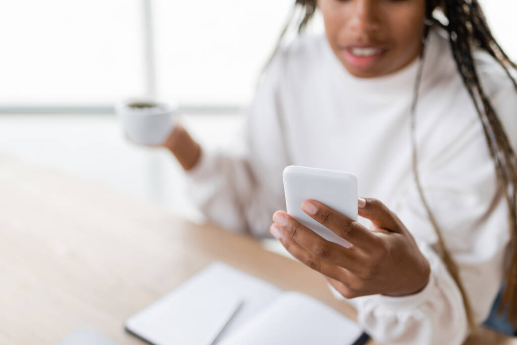Обрізаний вид розмитої афроамериканської бізнес-леді, використовуючи смартфон і тримаючи чашку в офісі
  - Фото, зображення