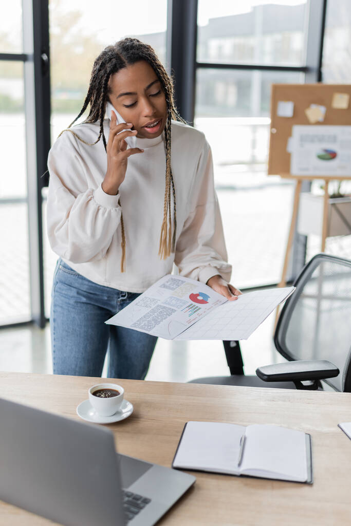 Afrykański amerykański bizneswoman rozmawia na smartfonie i trzyma papiery z wykresami w pobliżu kawy w biurze  - Zdjęcie, obraz