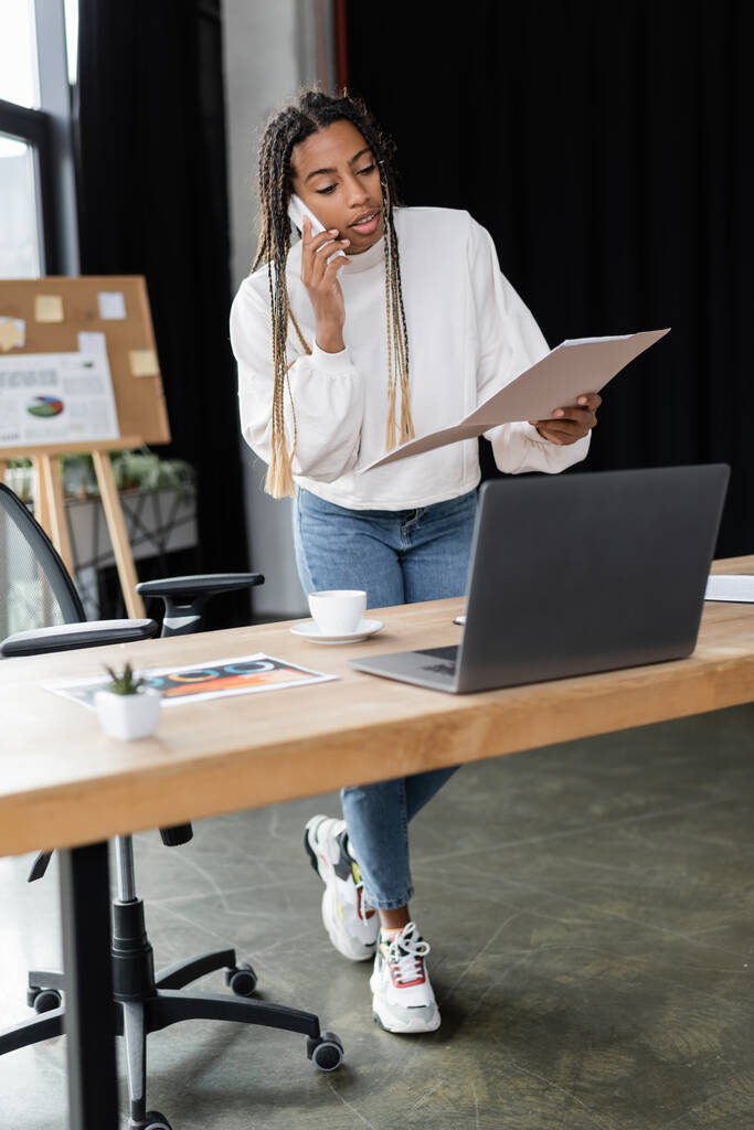 Afrikai amerikai üzletasszony beszél okostelefon és gazdaság papír mappa közelében laptop és kávé az irodában  - Fotó, kép