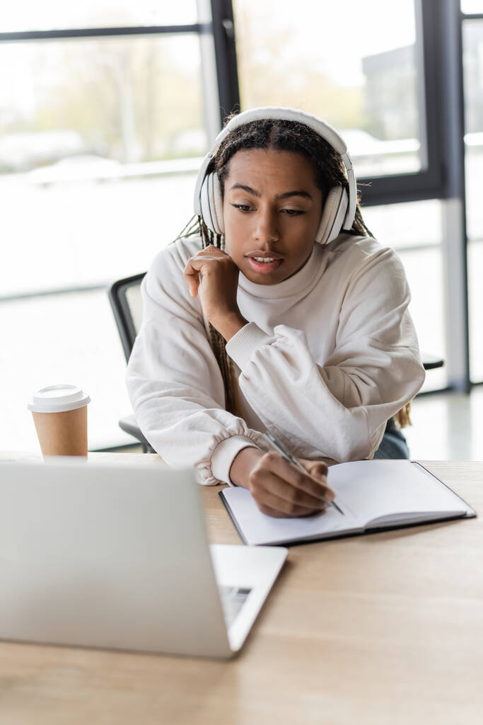 Mujer de negocios afroamericana en auriculares escribiendo en un cuaderno y mirando a la computadora portátil en la oficina  - Foto, imagen
