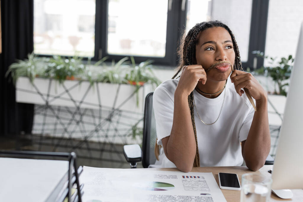 Pensiva mulher de negócios afro-americana tocando o cabelo perto de dispositivos no escritório  - Foto, Imagem