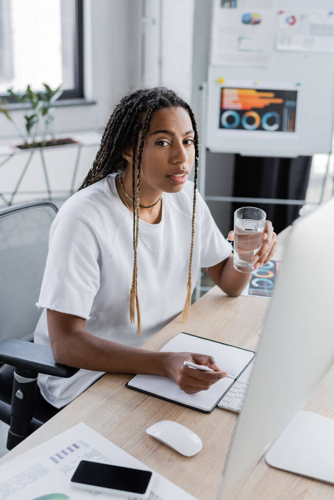 African American επιχειρηματίας κρατώντας ποτήρι νερό κοντά σημειωματάριο και υπολογιστή στο γραφείο  - Φωτογραφία, εικόνα
