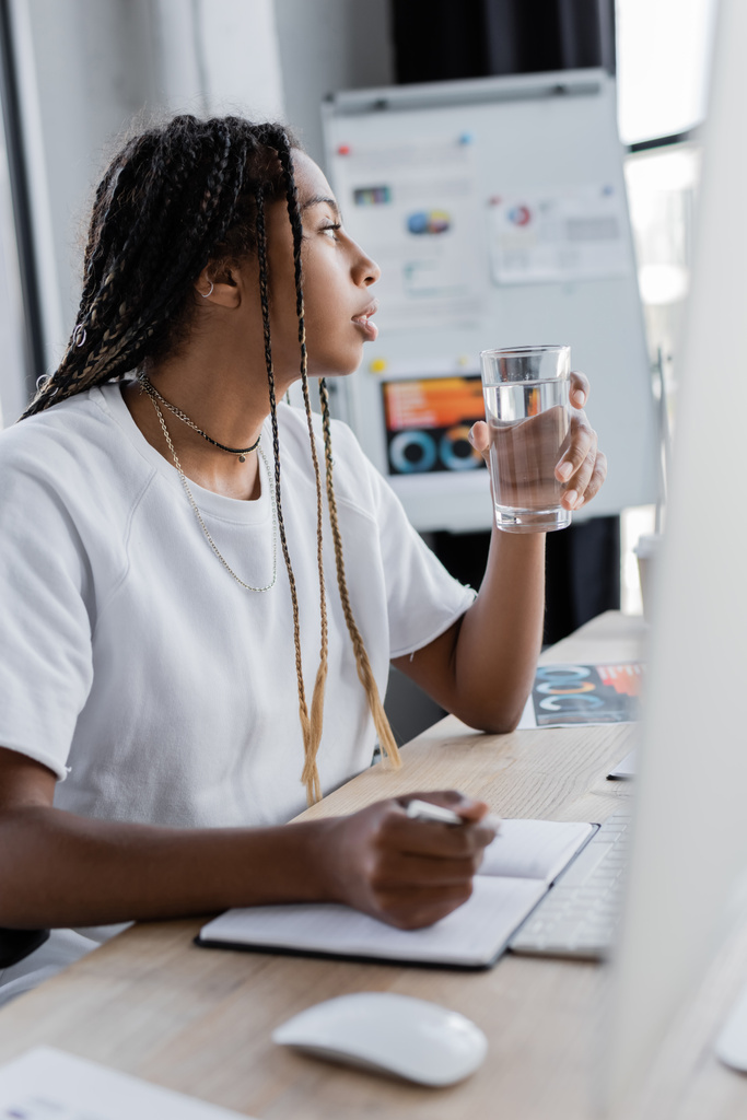 Vista laterale della donna d'affari afro-americana che tiene un bicchiere d'acqua vicino al notebook e al monitor del computer in ufficio  - Foto, immagini