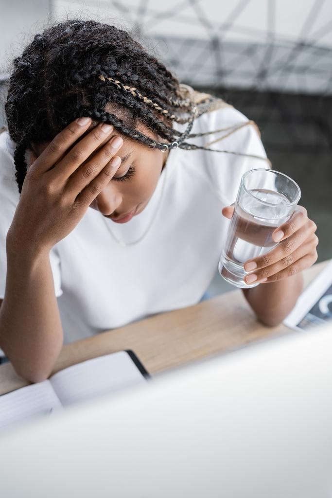 Vysoký úhel pohledu na unavený africký americký podnikatelek držící sklenici vody v blízkosti počítače v kanceláři  - Fotografie, Obrázek