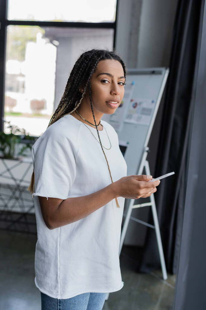Mujer de negocios afroamericana en ropa casual sosteniendo el teléfono inteligente y mirando a la cámara en la oficina  - Foto, imagen