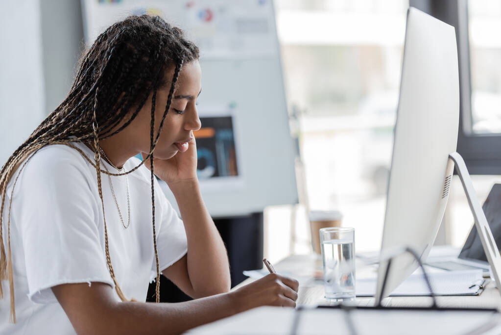 Afrykański amerykański bizneswoman w t-shirt pisania w pobliżu komputera i szklanki wody w biurze  - Zdjęcie, obraz