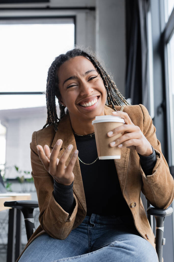 Позитивна афроамериканська комерсантка в куртці, яка тримає паперову чашку і дивиться на камеру в офісі  - Фото, зображення