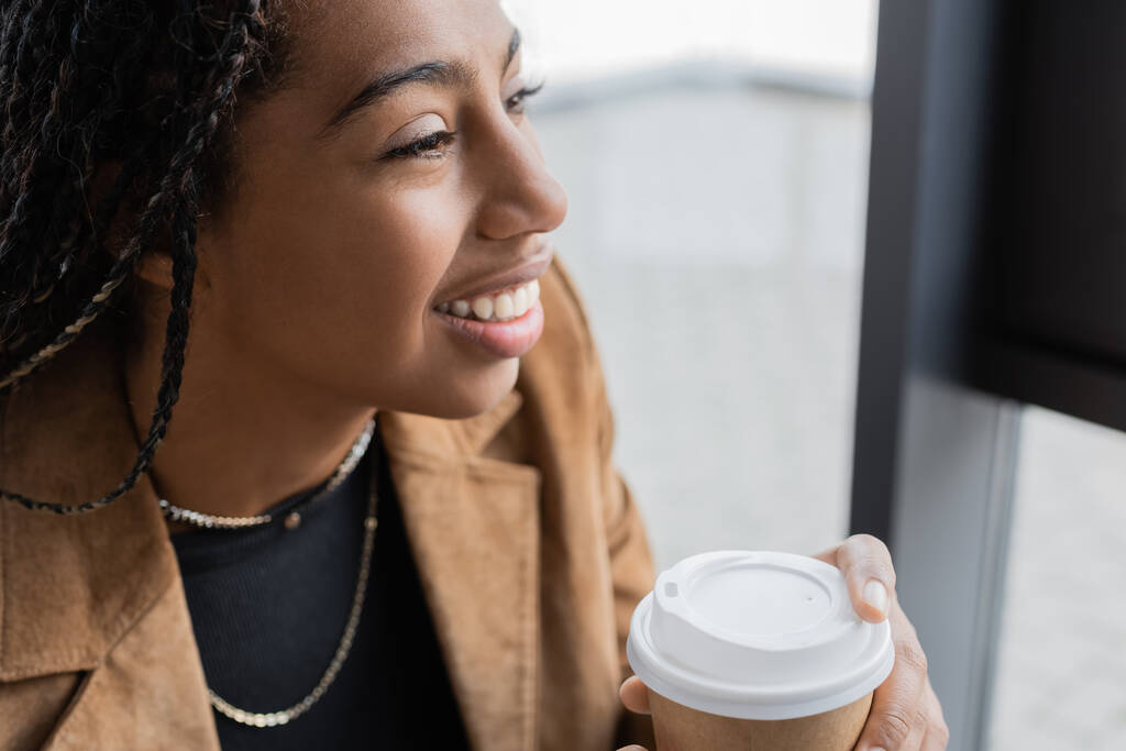 Sorridente donna d'affari afro-americana in giacca in possesso di tazza di carta in ufficio  - Foto, immagini