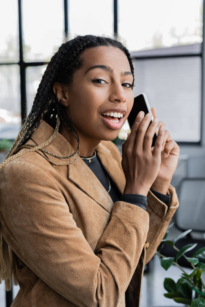 Femme d'affaires afro-américaine positive en veste tenant smartphone et regardant la caméra dans le bureau  - Photo, image