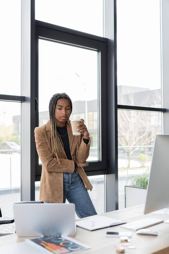 Donna d'affari afroamericana in possesso di caffè per avvicinarsi a dispositivi e notebook in ufficio  - Foto, immagini