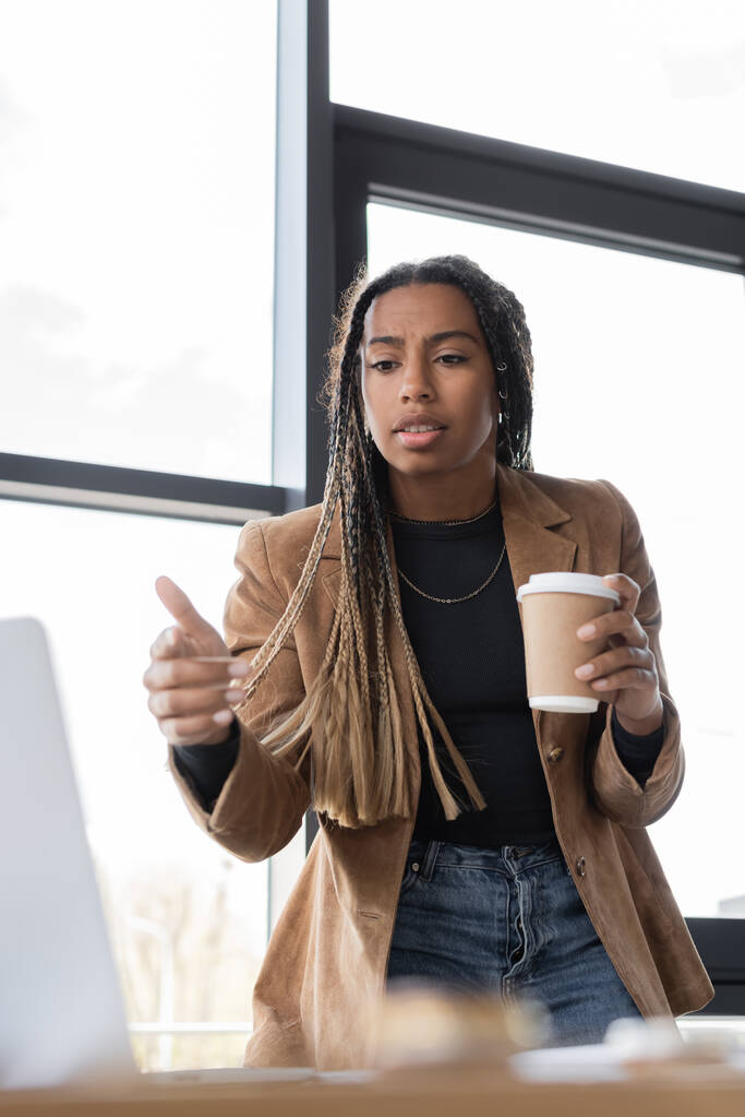 Donna d'affari afroamericana in possesso di caffè per andare e puntare con il dito al computer portatile in ufficio  - Foto, immagini