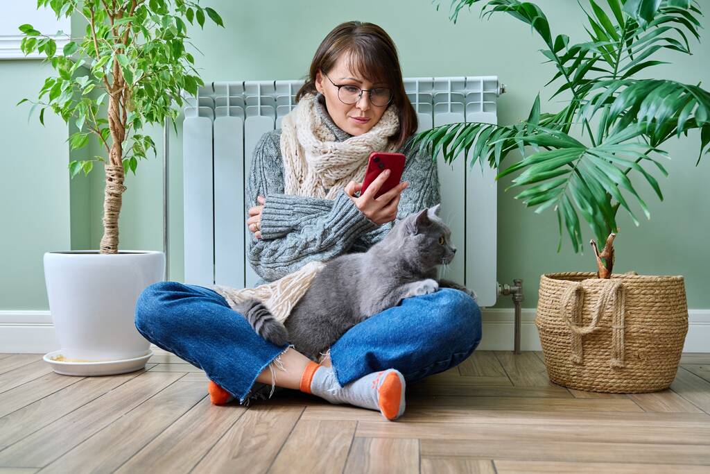 Middle aged woman resting at home in warm clothes using smartphone, warming herself with pet cat near home heating radiator. Winter autumn heating season, lifestyle leisure save energy, crisis concept - Zdjęcie, obraz