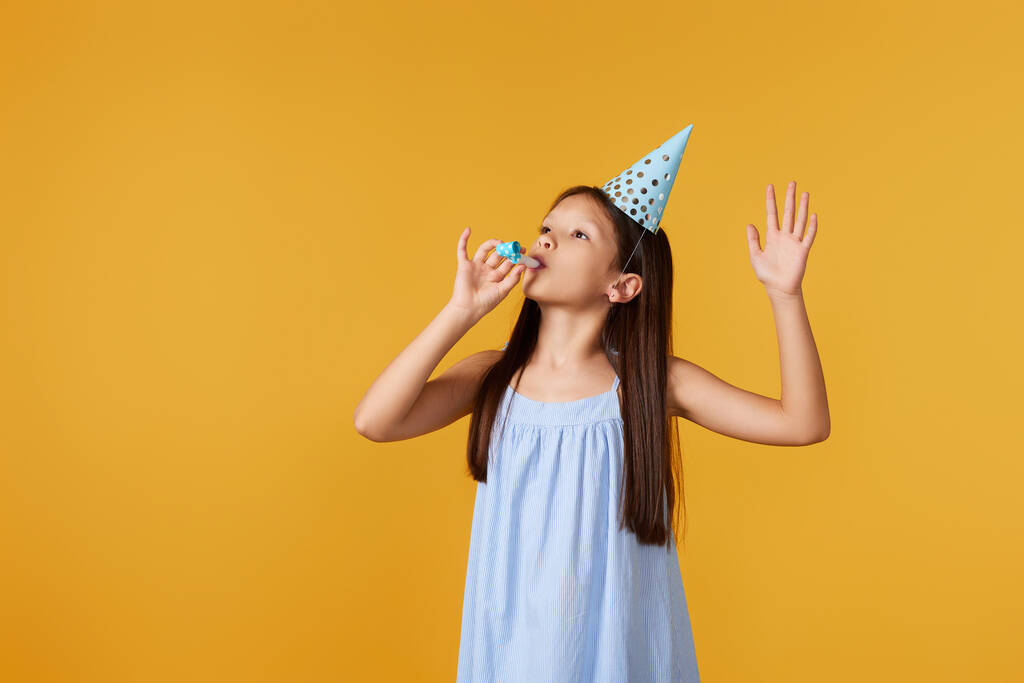 Parti konisi şapkalı ve sarı arka planda ıslık çalan mutlu küçük doğum günü kızı. Boşluğu kopyala - Fotoğraf, Görsel