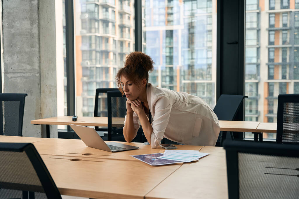 Ernsthaft fokussierte Geschäftsfrau lehnt am Schreibtisch und blickt auf den Laptop-Bildschirm - Foto, Bild