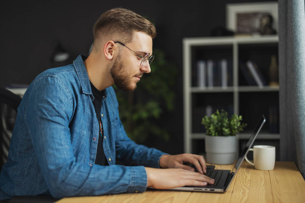 Joven hombre barbudo guapo escribiendo código en el ordenador portátil, programador con camisa de mezclilla y gafas en la oficina oscura, concepto - Foto, Imagen