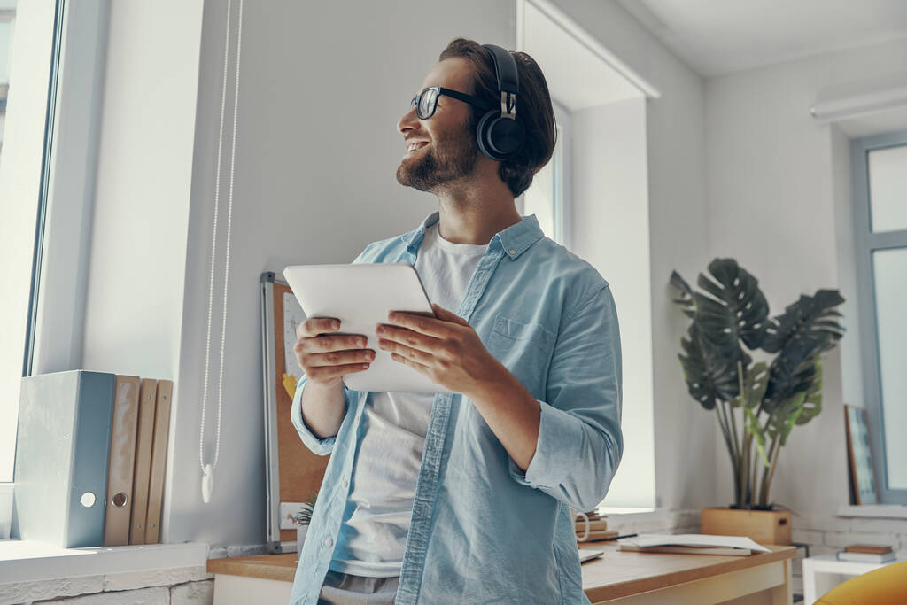 Ευτυχισμένος νεαρός άνδρας με ακουστικά κρατώντας ψηφιακό tablet, ενώ στέκεται στο γραφείο - Φωτογραφία, εικόνα