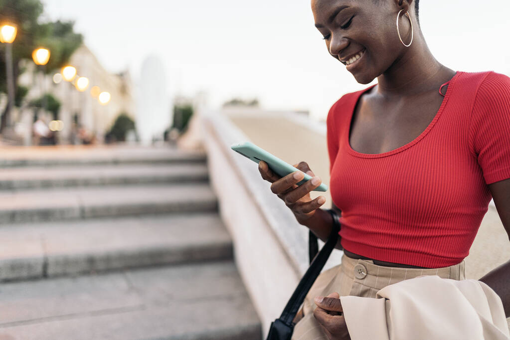 Detailní záběr na africké americké mladé dospělé ženy pomocí telefonu, zatímco drží její bundu a tašku na venkovních schodech ve městě - Fotografie, Obrázek