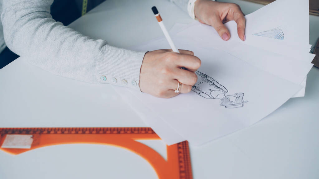 Close-up shot of young womans hand drawing outlines of stylish ladies garment with attention. Paper, tailors scissors, ruler are visible on studio table. Designer at work concept. - Photo, Image