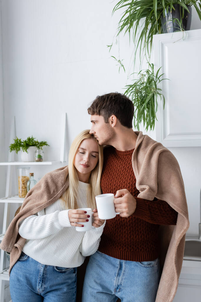 mladý pár pokrytý přikrývkou objímání při držení šálky čaje doma - Fotografie, Obrázek