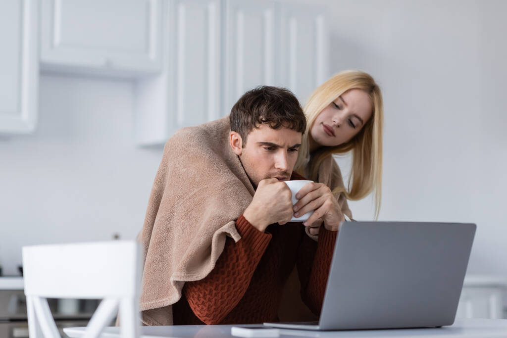 blonde woman holding blanket near boyfriend with cup working remotely from home - Photo, Image