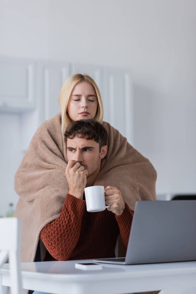 mulher loira segurando cobertor perto namorado preocupado com copo trabalhando remotamente de casa - Foto, Imagem