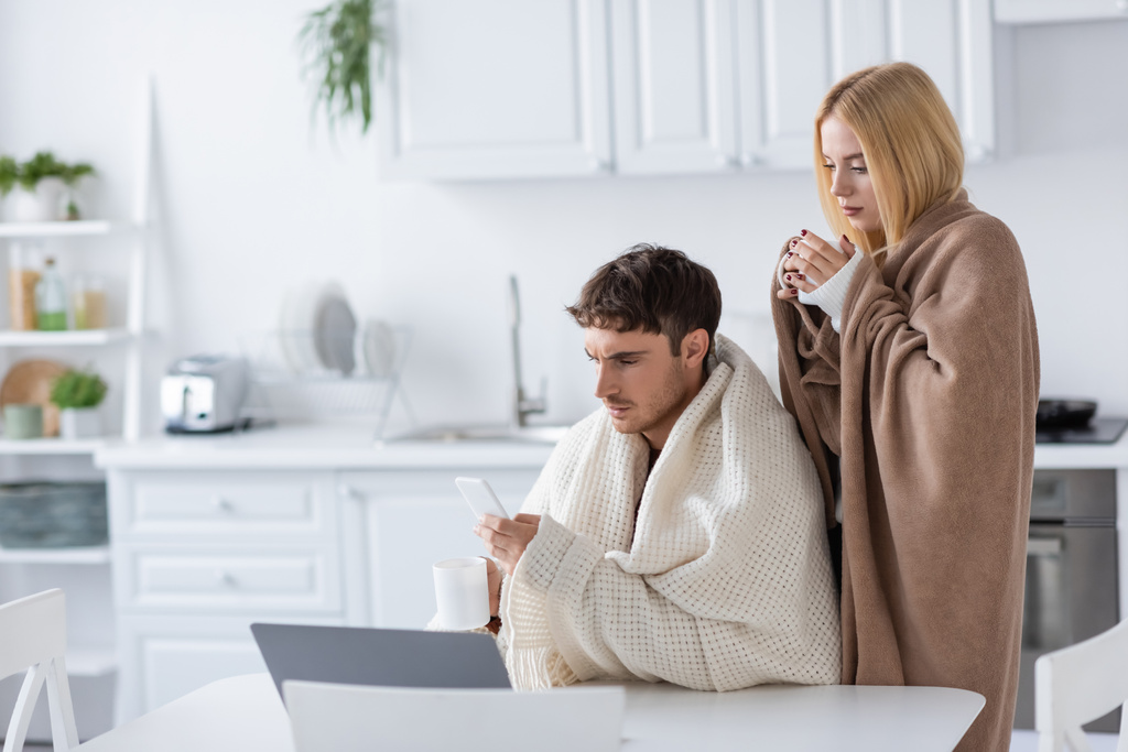 femme blonde couverte de couverture près de petit ami avec smartphone et tasse  - Photo, image
