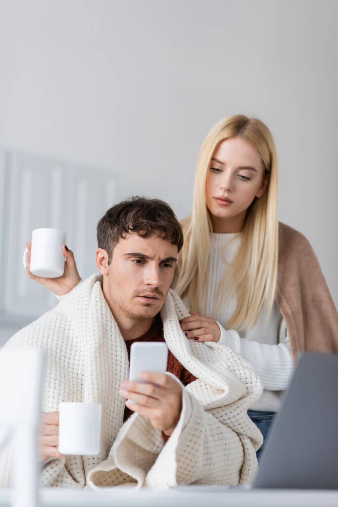 young woman covered in blanket standing near boyfriend with smartphone and cup of tea - Photo, Image