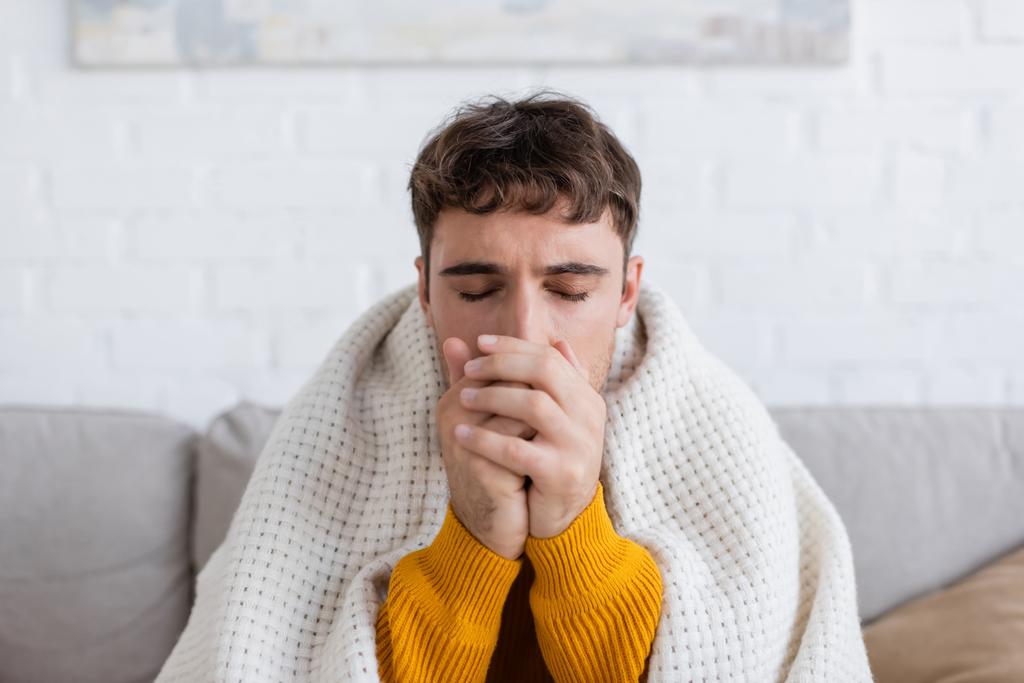 junger Mann mit geschlossenen Augen sitzt in Decke gehüllt und wärmenden Händen im Wohnzimmer  - Foto, Bild