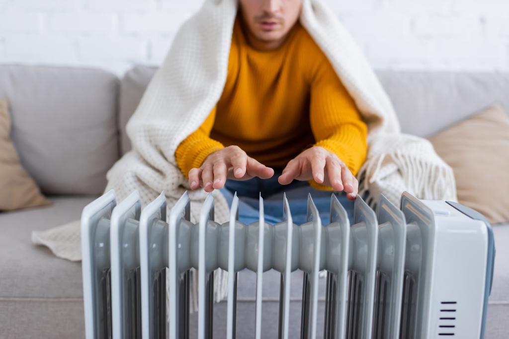 обрезанный вид молодого человека, покрытого одеялом, сидящего на диване и согревающегося у обогревателя радиатора зимой  - Фото, изображение