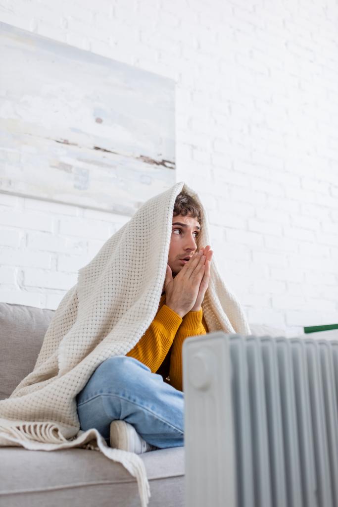 jonge man bedekt met deken zitten op de bank en opwarmen van handen in de buurt radiator verwarming - Foto, afbeelding