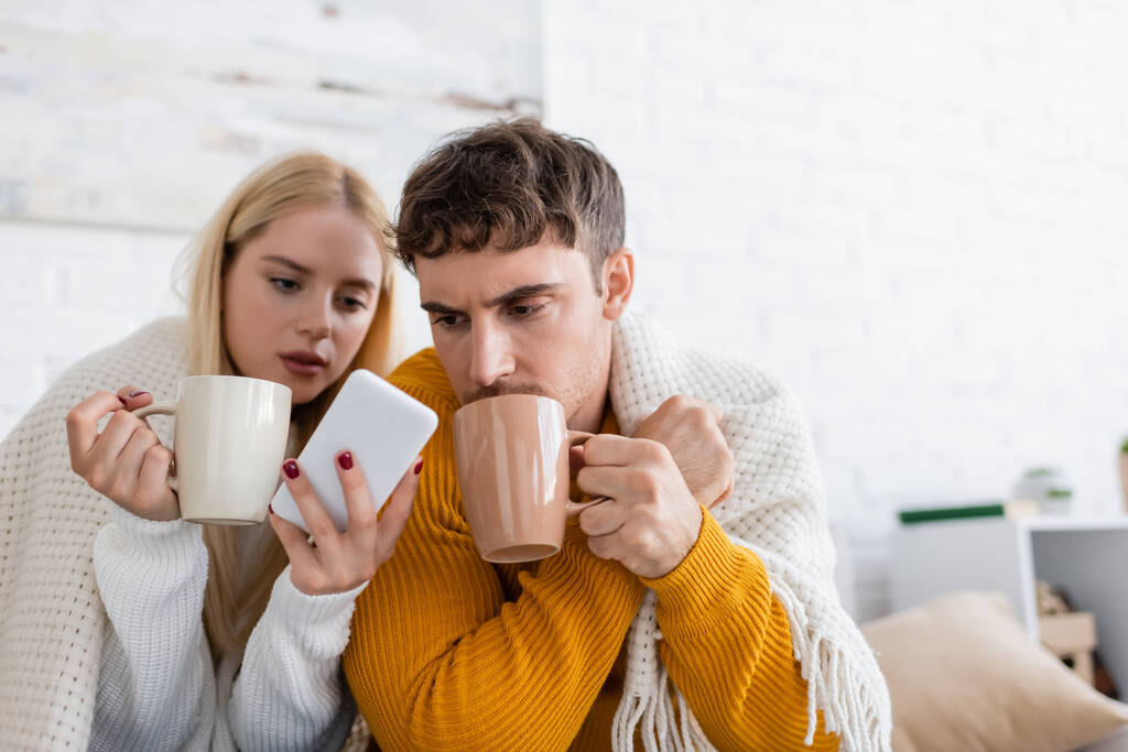 Elinde çay ve akıllı telefon olan genç bir kadın battaniyeyle erkek arkadaşının yanında.  - Fotoğraf, Görsel