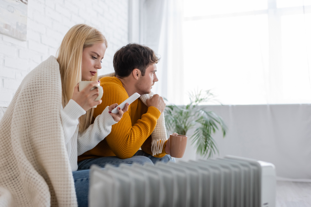 jeune femme avec tasse de thé à l'aide d'un smartphone près du petit ami recouvert d'une couverture tout en étant assis près du radiateur - Photo, image