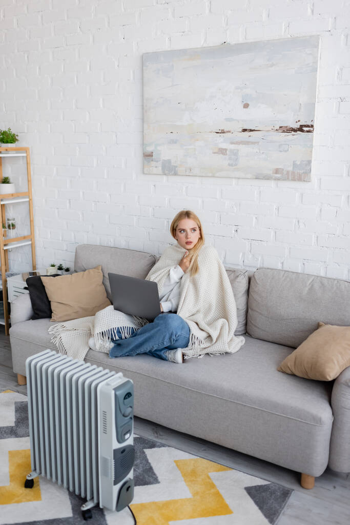 jeune femme blonde recouverte d'une couverture blanche à l'aide d'un ordinateur portable assis sur un canapé près du radiateur - Photo, image