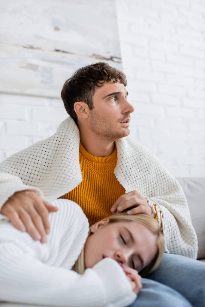 blonde woman covered in blanket sleeping on legs of boyfriend in jeans sitting on couch - Photo, Image