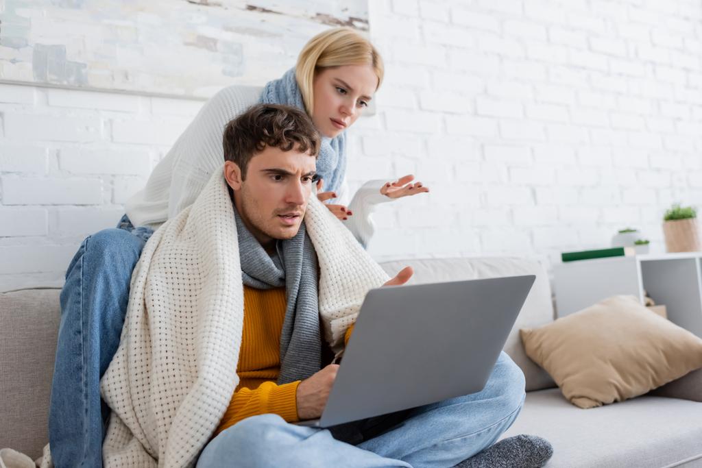 niezadowolona blondynka w swetrze i szaliku siedzi obok chłopaka i gestykuluje patrząc na laptopa  - Zdjęcie, obraz