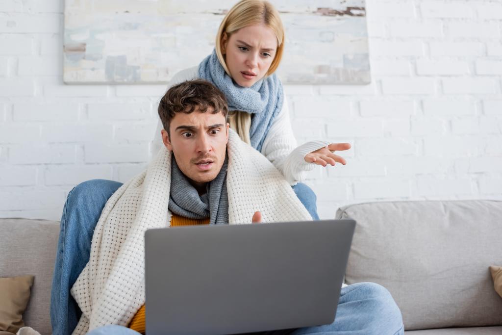 mulher loira descontente em suéter e cachecol sentado perto namorado chocado e gestos enquanto olha para laptop  - Foto, Imagem