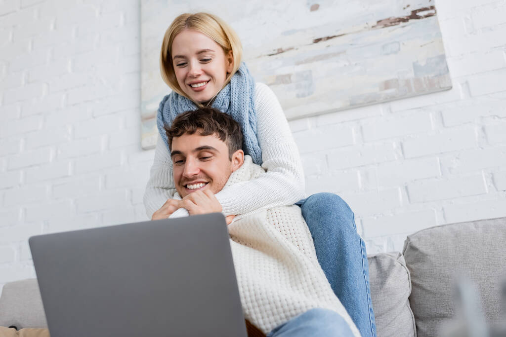 mujer rubia feliz en suéter y bufanda abrazando novio alegre mientras mira el ordenador portátil  - Foto, imagen
