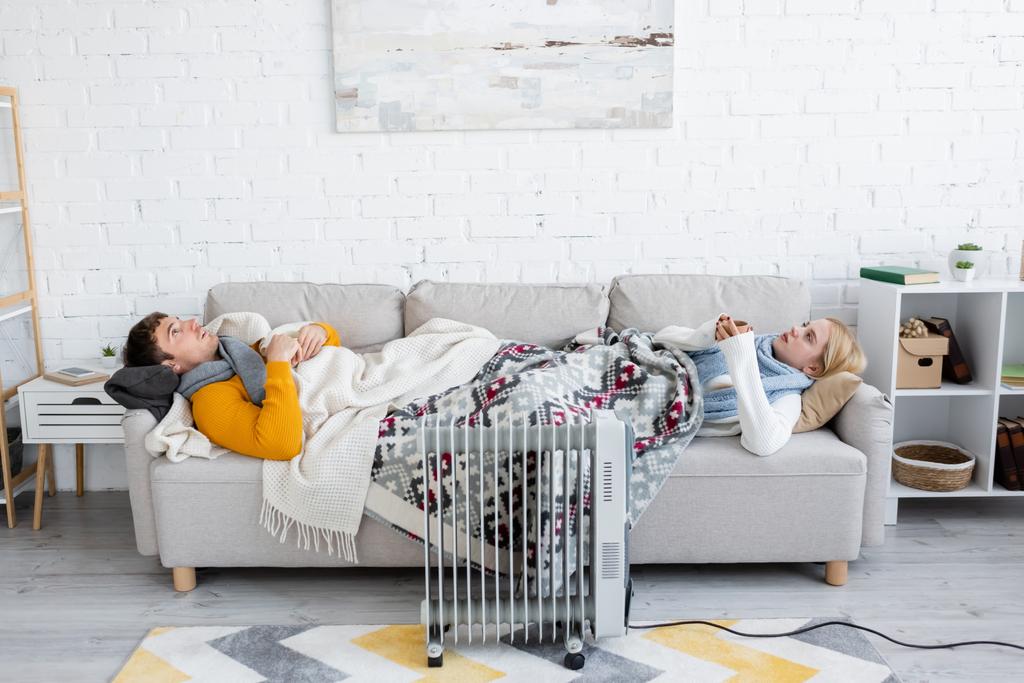 young man and blonde woman holding cups and lying under blankets on sofa near radiator heater - Photo, Image