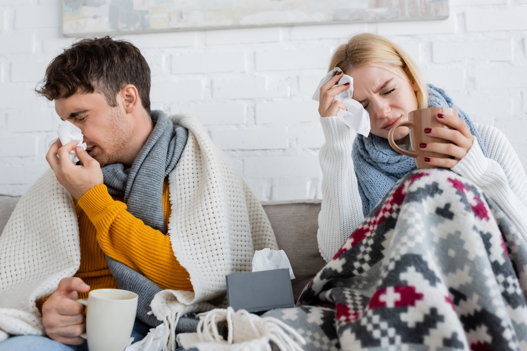 Erkrankter Mann niest neben blonder Frau mit Migräne und hält Tasse Tee  - Foto, Bild
